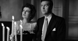 A woman and a man in formal attire stand near lit candles, looking off to the side in a black-and-white photograph.