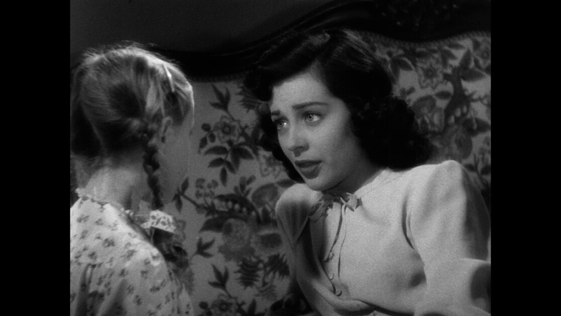 A black-and-white scene showing a woman with dark hair speaking to a child with blonde braided hair. They are sitting on a patterned couch.