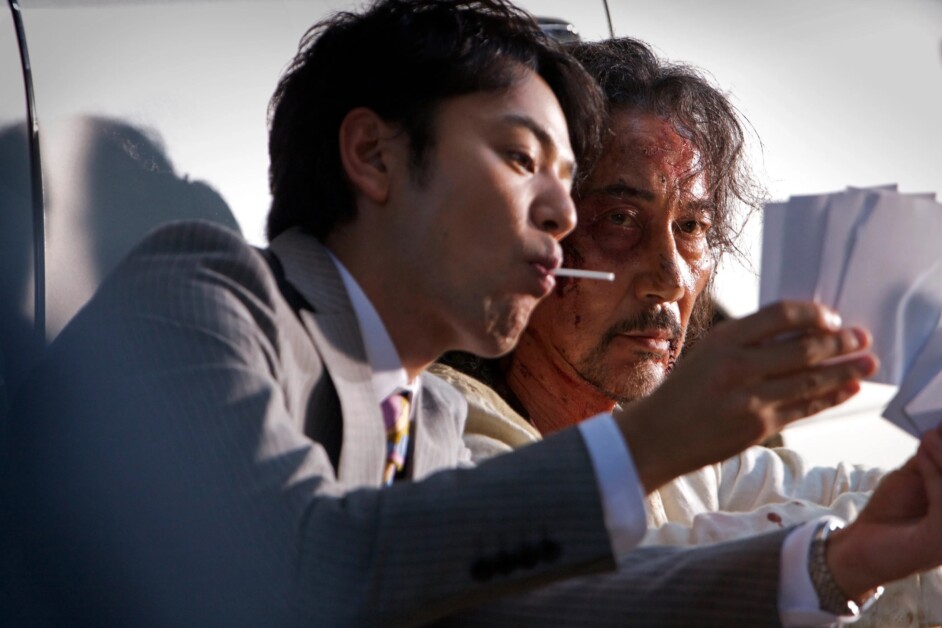 A man in a suit with a lollipop reads documents while an older man with bloodstains on his face looks on.