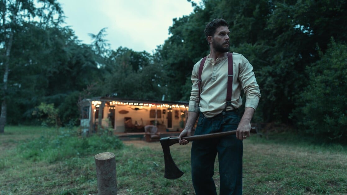 A man with a beard holds an axe near a chopped tree stump. He stands in front of a house with string lights in a wooded area.