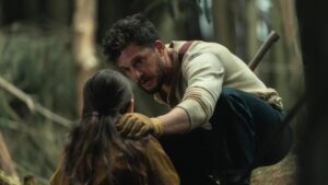 A man crouches in a forest, talking to a young girl with long hair. He is wearing a white shirt, gloves, and has an axe strapped to his back.