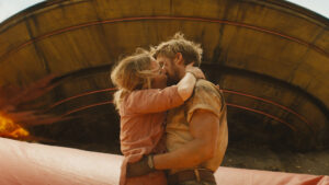 A man and woman embrace and kiss in front of a large, round, metallic structure. Both are dressed in casual, rugged clothing.