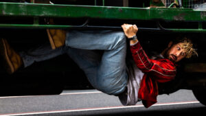 A person in a red jacket and jeans hangs underneath a vehicle, gripping a metal bar with both hands.