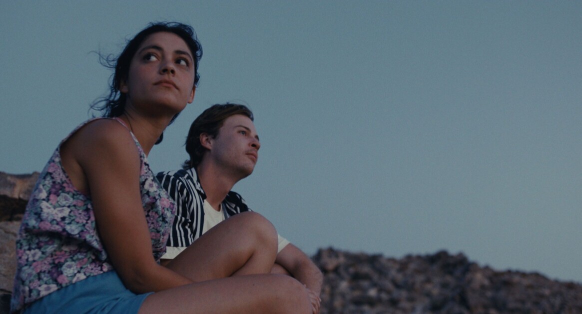 Two individuals are seated outdoors, one wearing a floral top and shorts, and the other in a striped shirt. They appear to be gazing into the distance with a rocky landscape in the background.