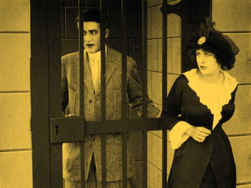 A man stands behind bars while a woman in a period dress stands outside, looking concerned. The image has a yellow tint.