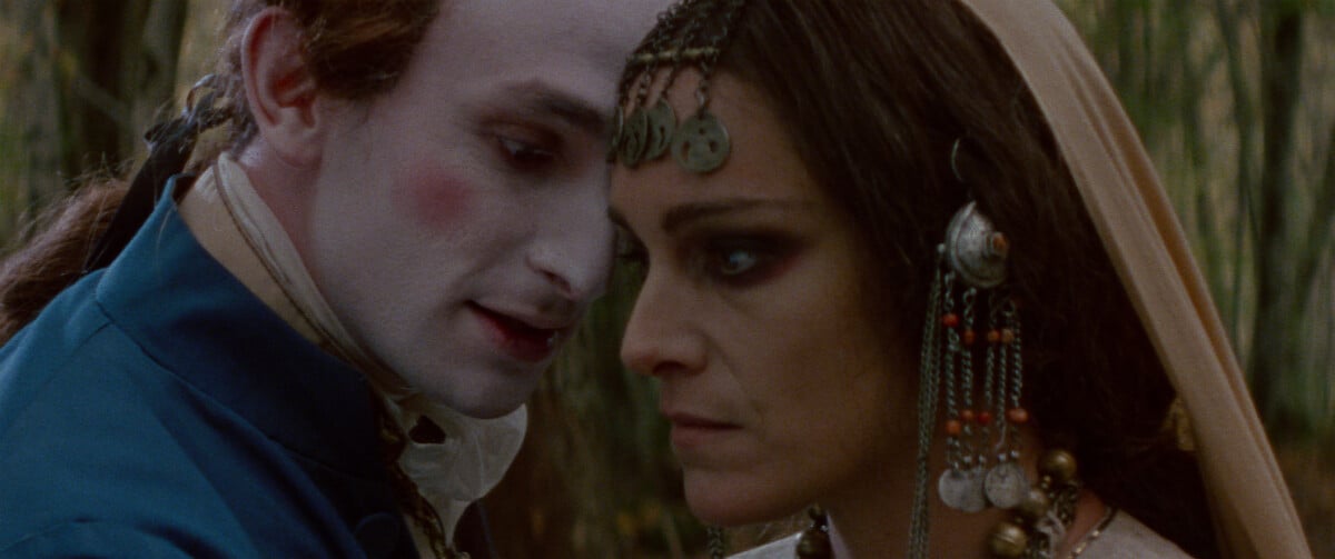 A person with a painted white face and elaborate costume leans close to a person with heavy eye makeup and decorated headscarf, set in an outdoor environment.