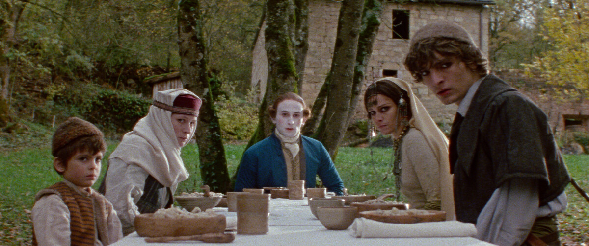 A group of people in period costumes sit at a long outdoor dining table, with neutral expressions. Behind them, trees and a stone building are visible.