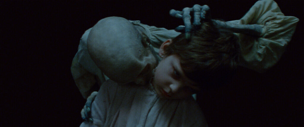 A figure with a skeletal appearance touches a child's head while the child looks down, both set against a dark background.