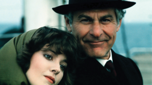 A woman with short brown hair rests her head on the shoulder of an older man wearing a hat. Both are looking at the camera, and the background is blurred.