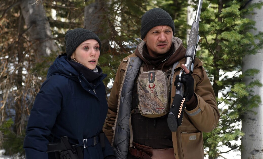 Two people dressed in winter clothing stand in a snowy forest. One holds a shotgun, and both appear alert.