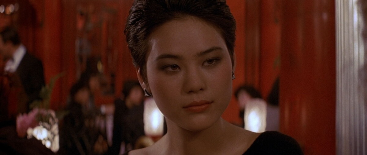 A woman with short, dark hair and an expressionless face in a dimly lit, red-toned setting, possibly a restaurant. People and decor are visible in the background.