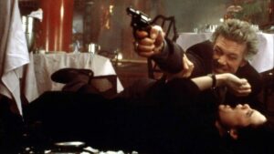 A man holding a gun lies on top of a woman on the floor of what appears to be a restaurant, with tables and red decor visible in the background.