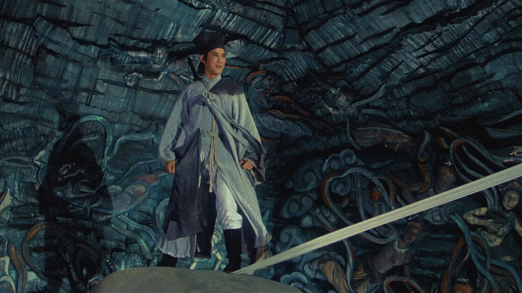A person wearing traditional clothing stands on a rock with intricate, mythological artwork on the cave walls in the background.
