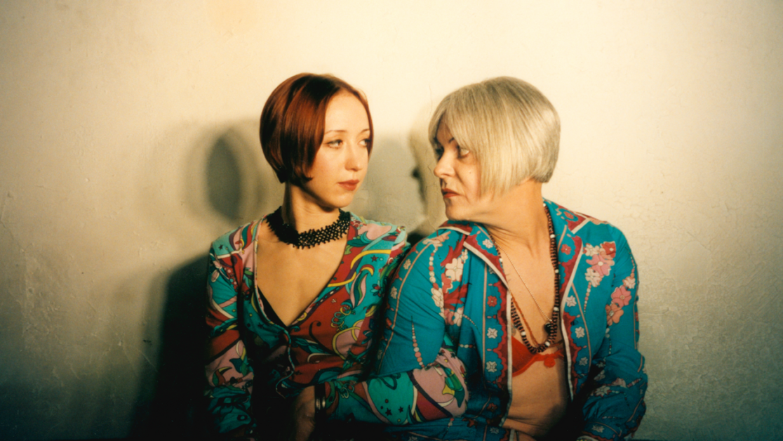 Two individuals with short haircuts and colorful, patterned outfits sit side by side against a plain background, looking in different directions.