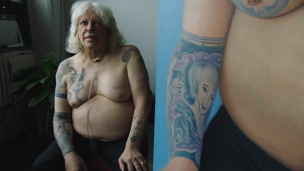 A shirtless person with tattoos on their upper body sits near a window. The right side of the image shows a close-up of an arm tattoo featuring a face and text.
