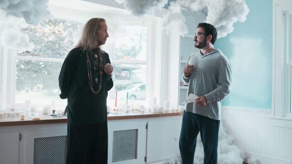 Two men stand in a brightly lit room surrounded by artificial clouds. The man on the left wears a black outfit with beaded necklaces, while the man on the right sports glasses and holds papers.