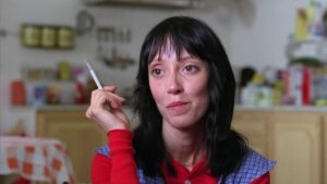 A person with black hair holds a cigarette while sitting in a kitchen. Various kitchen items and a checkered tablecloth are visible in the background.