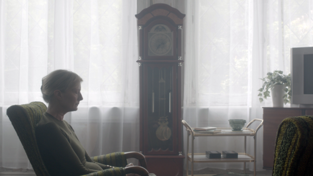 A person sits in a green armchair facing sideways in a dimly lit room with a tall grandfather clock, a cart with cups and books, and a television. White curtains cover the large windows in the background.