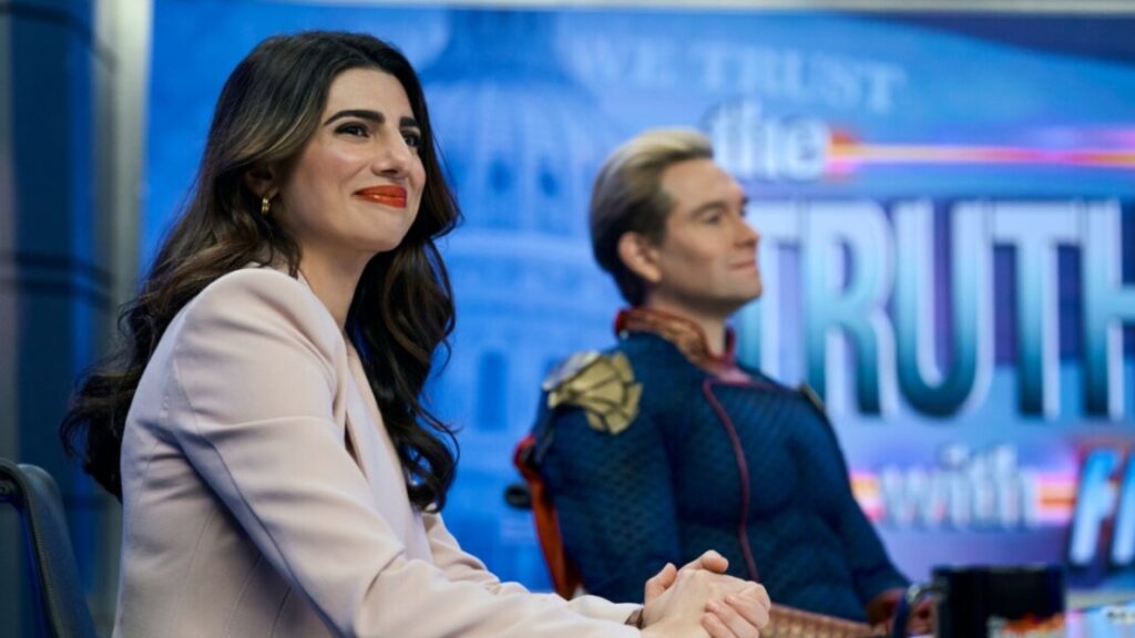 A woman in a light-colored blazer and a man in a superhero outfit are seated side by side, smiling, in a studio with "THE TRUTH" displayed in the background.