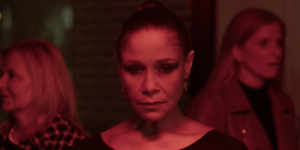 A person with a serious expression stands under red lighting, surrounded by others in the background.
