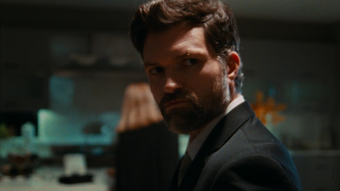 A bearded man in a suit stands in a dimly lit room, looking over his shoulder. A blurred figure in the background appears to be walking away.