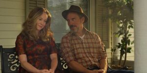 Inspiring Drama ‘A Carpenter’s Prayer’ Premieres Nationwide On August 9 A man and a woman sit on a porch bench, engaged in conversation. The woman wears a patterned dress, and the man wears a plaid shirt and a hat. A potted plant is visible next to them.