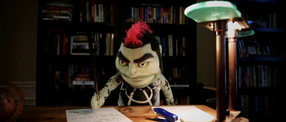 A puppet with a pink mohawk and black clothes writes on paper at a desk with a lamp, in a room with bookshelves in the background.