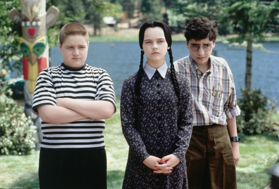 Three young people standing outdoors near a lake with trees in the background. One wears a striped shirt, one wears a floral dress, and one wears a plaid shirt with a pocket protector.