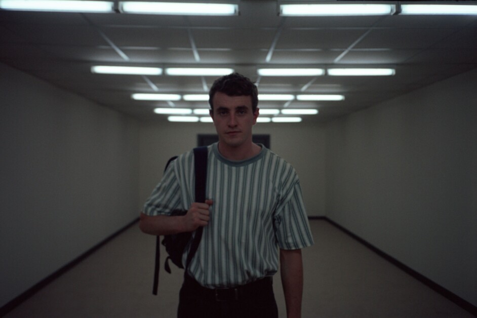 A person wearing a striped shirt and carrying a backpack walks down a hallway illuminated by overhead fluorescent lights.