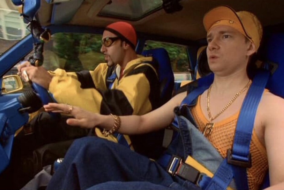 Two people are in a car. The driver wears a yellow jacket and cap. The passenger wears a yellow bandana, orange mesh top, and overalls, looking concerned with a hand extended towards the dashboard.