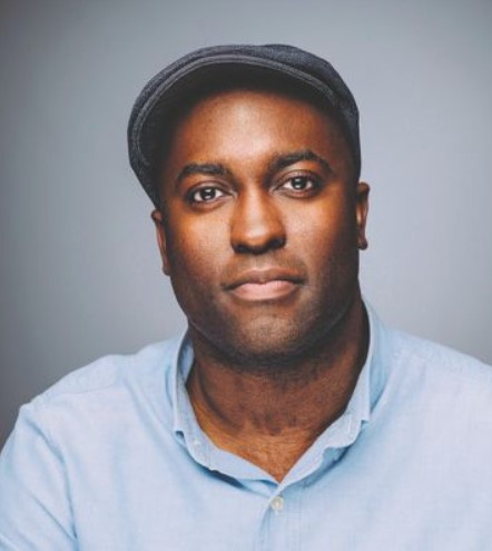 A person wearing a flat cap and a light blue button-up shirt poses against a plain gray background.
