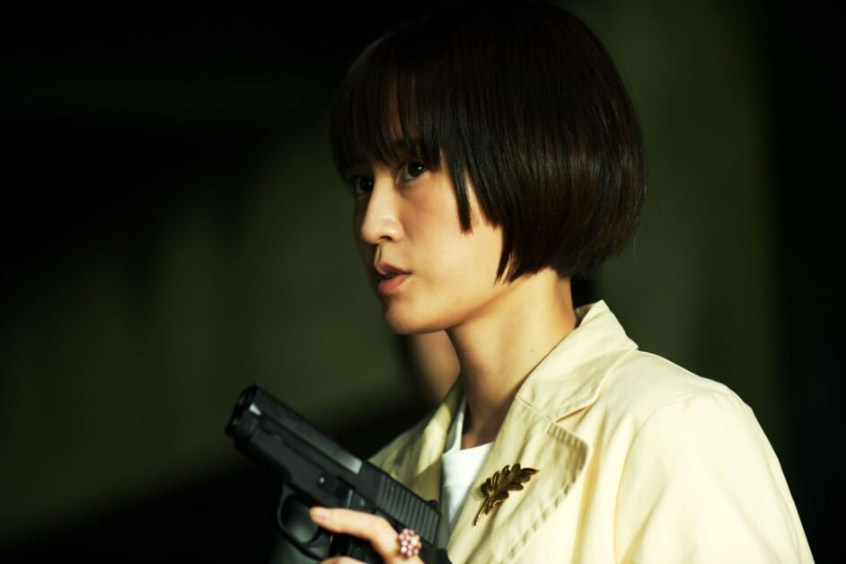 A person with short, dark hair wearing a light-colored jacket holds a black handgun.