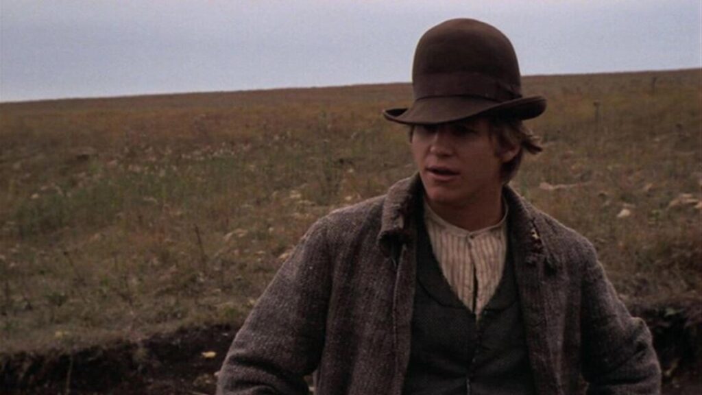 A person wearing a brown hat and a grey coat stands outdoors in a field with a cloudy sky in the background.