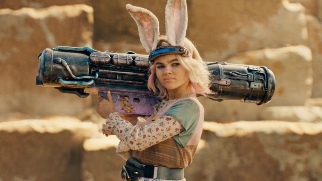 Person with bunny ears headband holding a large futuristic weapon, wearing a floral-patterned top and a tactical vest, standing against a rocky background.