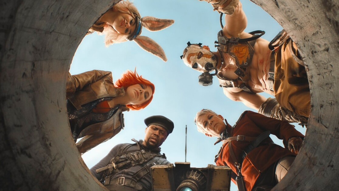 Five people in costumed attire look down into a circular opening from above, against a clear sky background.