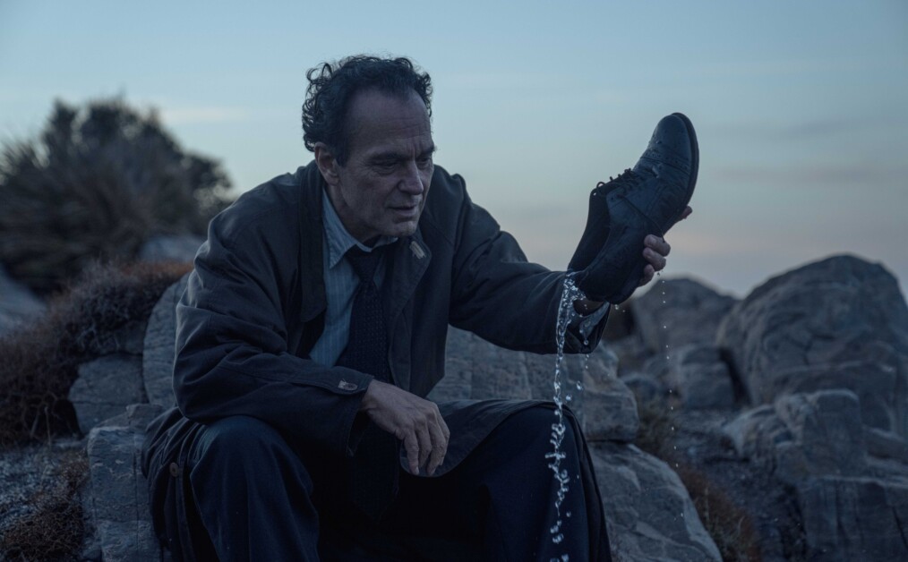 A man sits on rocks outdoors, holding up a dress shoe with water pouring out of it, looking thoughtful.