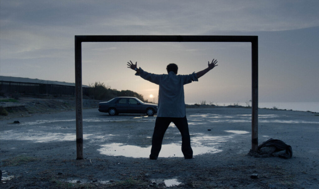 A person with arms outstretched stands in an empty goalpost on a deserted, wet beach, with a parked car and the ocean in the background at sunset.