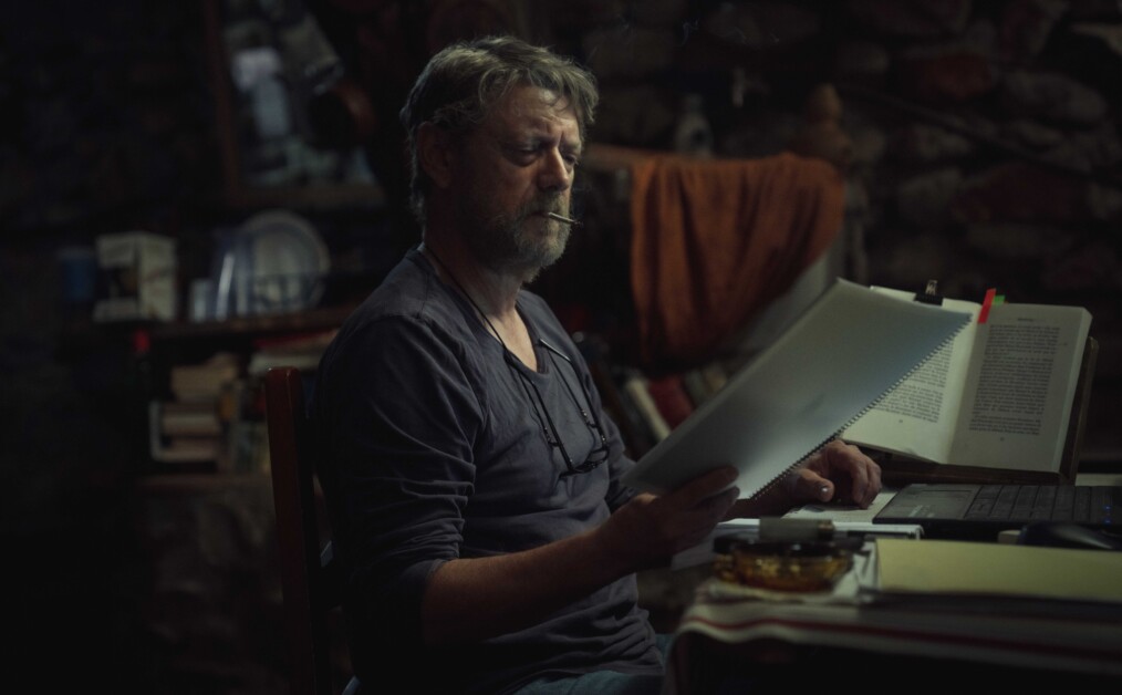A man with gray hair and a beard is sitting at a table, looking at a document. There is a computer, an ashtray, and books in the dimly lit room.