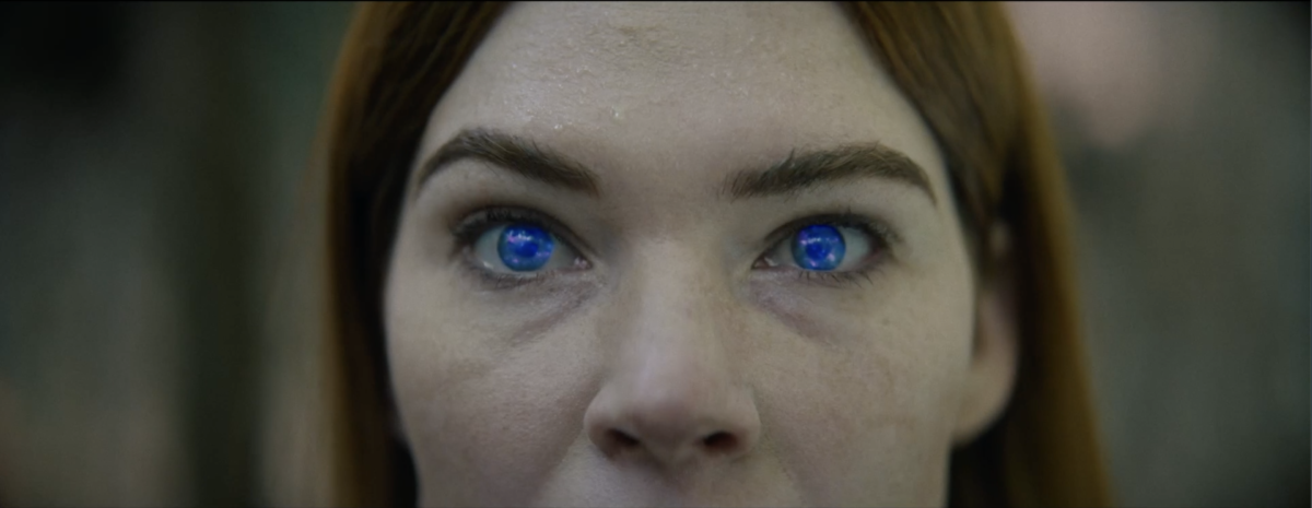 Close-up of a person's face with blue glowing eyes and a focused expression. The background is blurred.