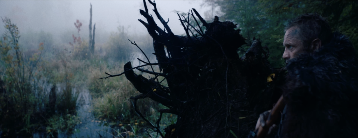 A person in fur outerwear stands partially obscured behind a large, gnarled tree stump in a foggy, dense forest setting.