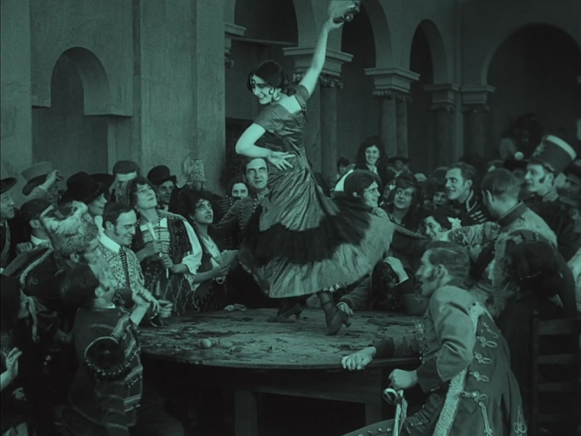 A woman in a dress dances with one arm raised on a table surrounded by a crowd of people watching cheerfully in an indoor setting.