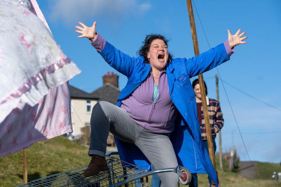 A person in a blue coat stands excitedly with arms outstretched, one leg resting on a shopping cart in an outdoor setting. Another person is seen in the background.