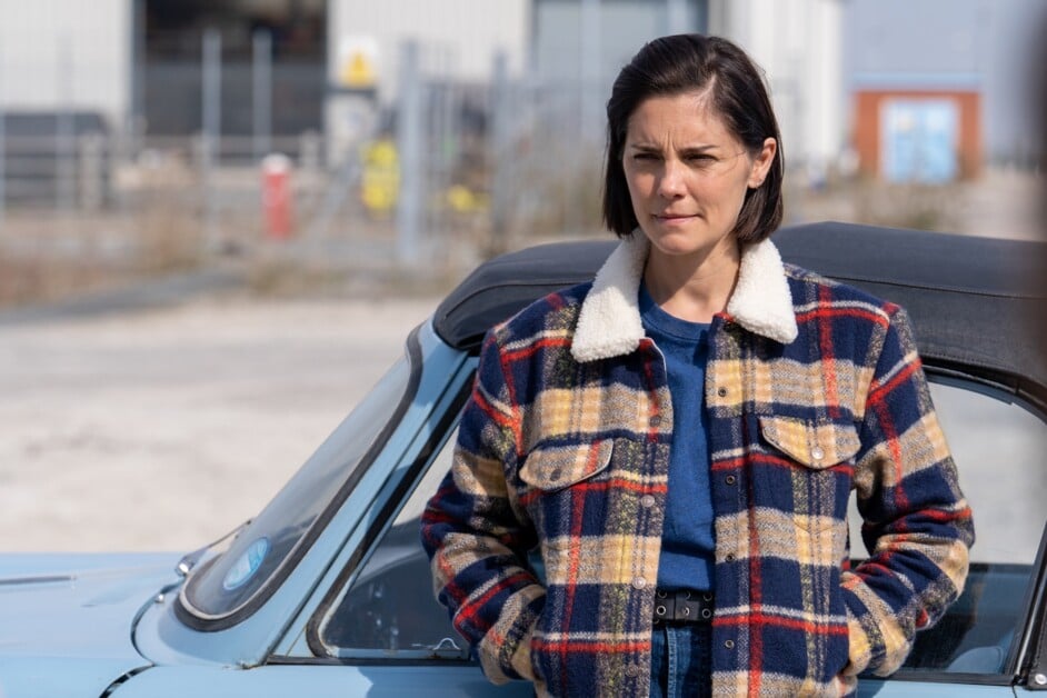 A woman with short dark hair wearing a plaid jacket stands next to a light blue car in an outdoor setting.