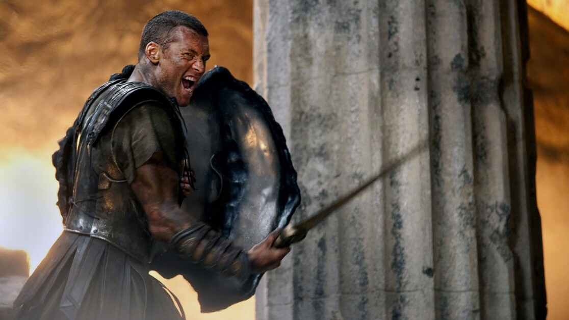 A man in ancient armor, holding a large shield and a sword, is shouting fiercely in front of stone columns.