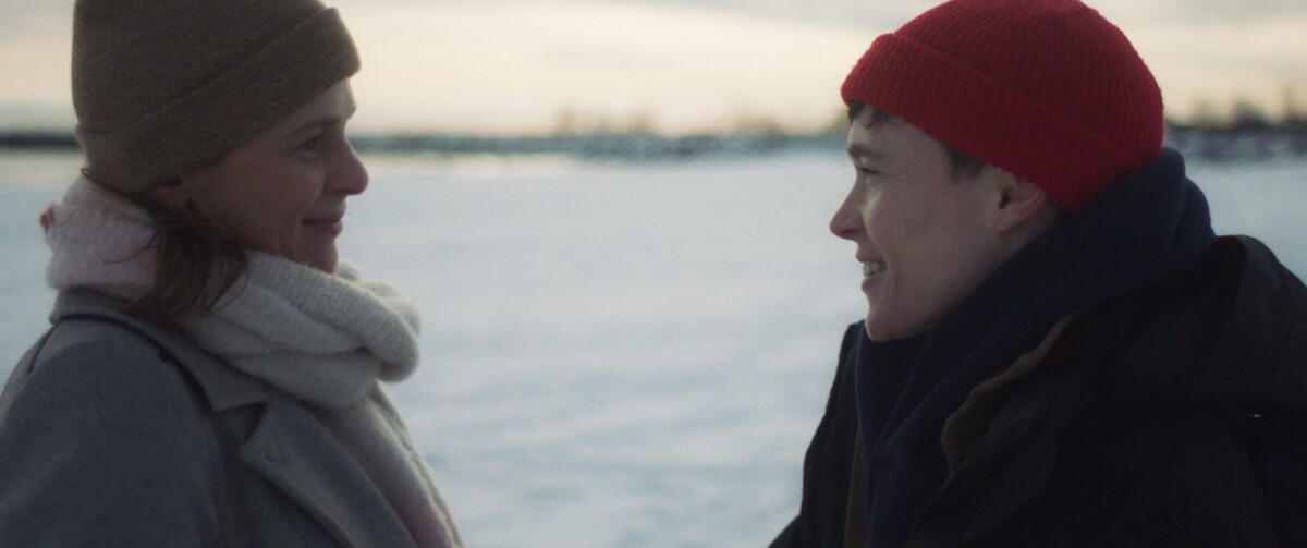Two people wearing winter clothes, including hats and scarves, stand facing each other and smiling in a snowy outdoor setting.