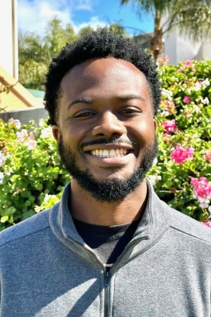 A man with a beard and short curly hair smiles at the camera, standing in front of a background filled with green foliage and blooming pink flowers.