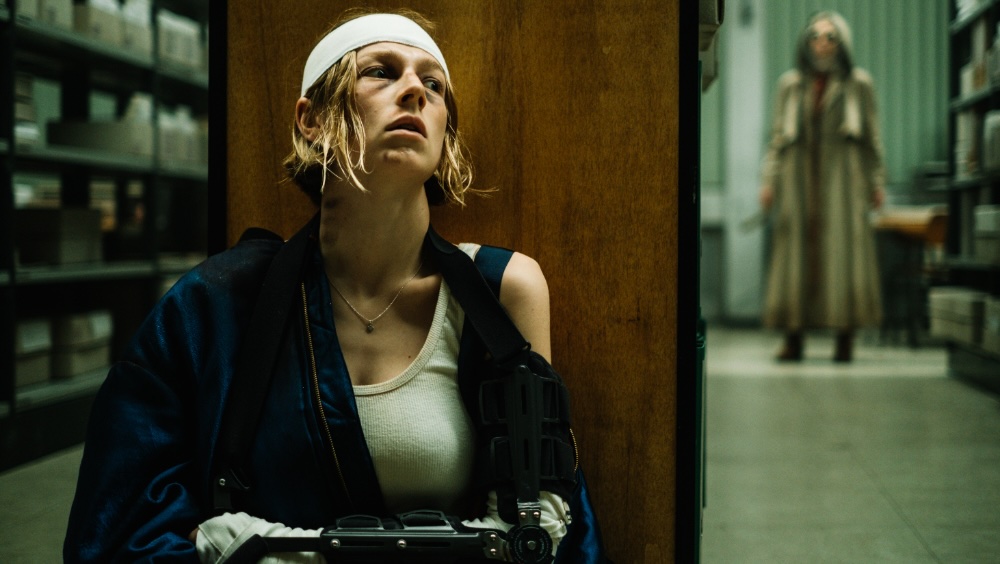 A person with a head bandage and arm brace sits against a wooden panel in what appears to be a storage area, while another person in a long coat stands in the background.