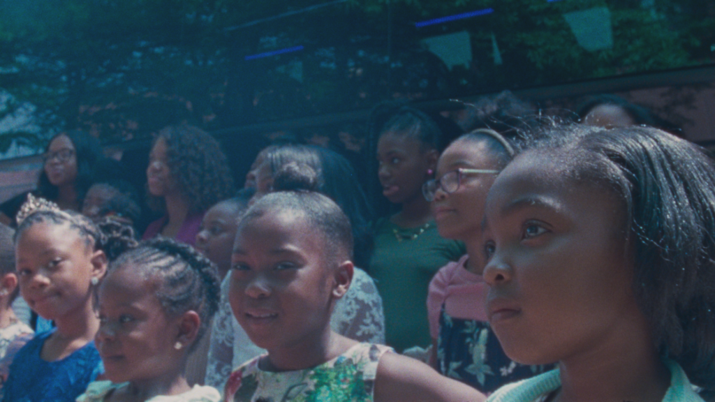 A group of children and adults, all dressed in colorful clothing, stand closely together outdoors, looking towards something off camera.