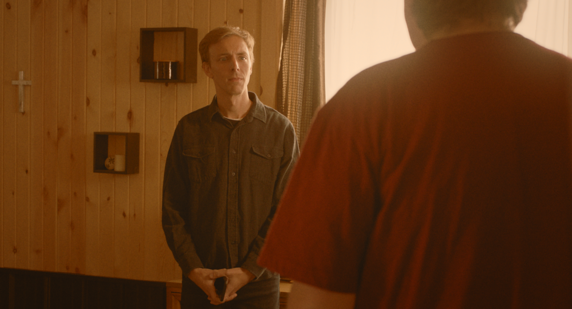 A man in a gray shirt stands facing another man in a red shirt in a wooden-paneled room with a cross and small shelves on the wall.