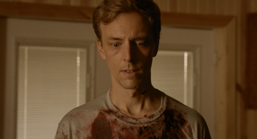 A man with disheveled hair and a distressed expression stands indoors wearing a white T-shirt stained with blood.